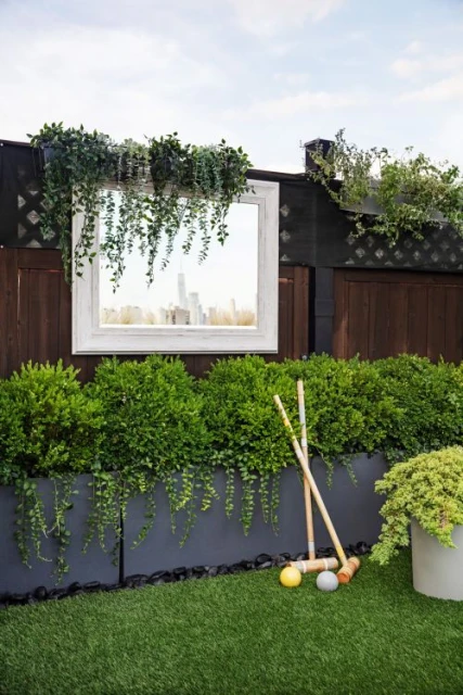 Croquet set and greenery in an outdoor setting with a framed mirror reflecting a cityscape, evokes a blend of nature and urbanity.