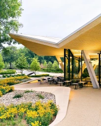 Modern restaurant exterior with a sweeping folded roof, glass walls, outdoor patio seating, and surrounding flower beds.