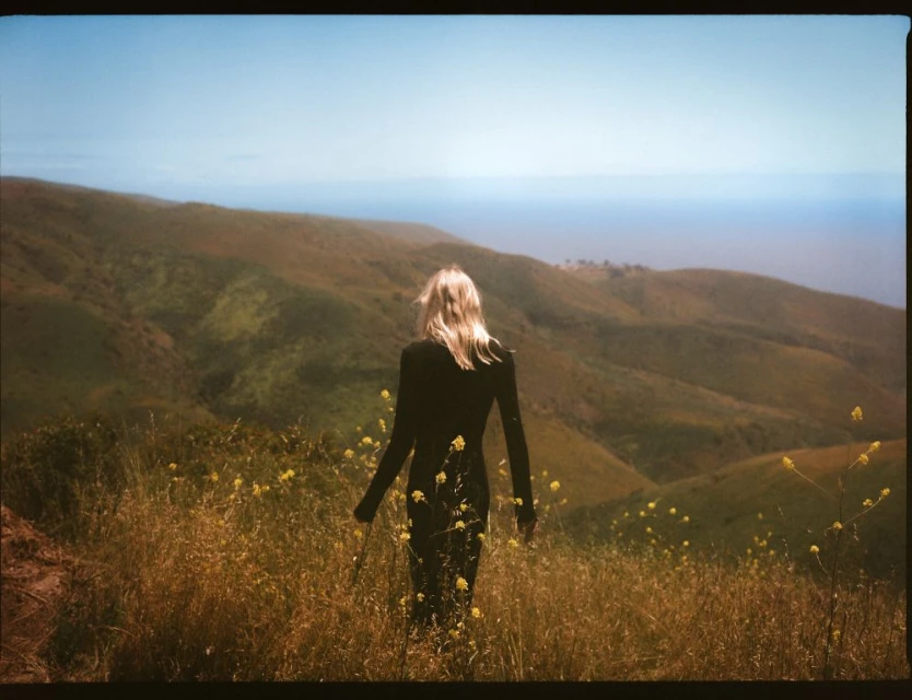Blonde woman in black dress walking through golden field with yellow wildflowers, rolling green hills, and distant ocean on the horizon.