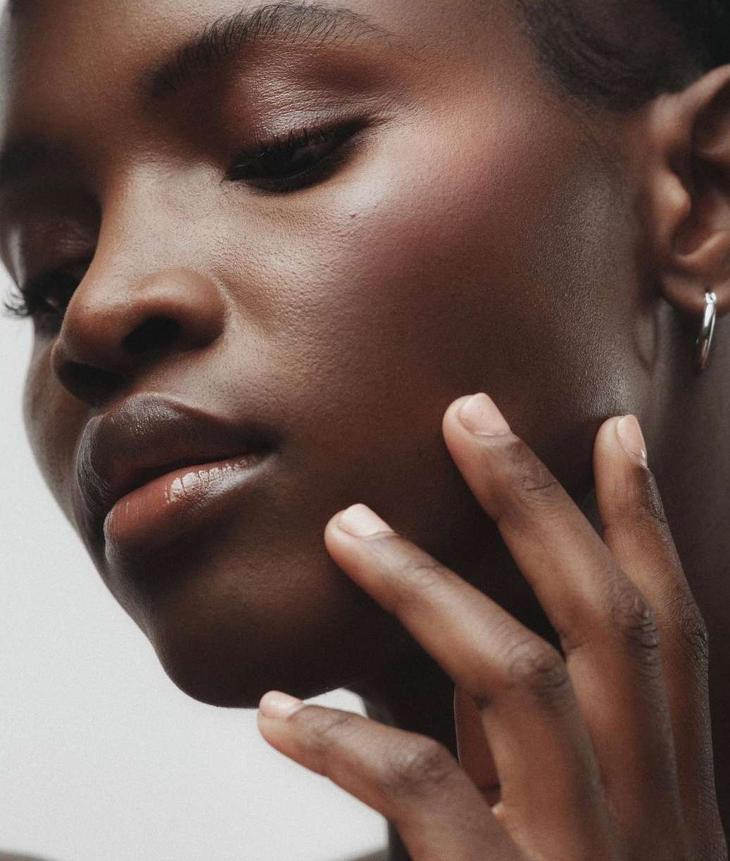 Close-up studio shot of Black woman touching her face. Beauty portrait features skin detail, minimal makeup, silver hoop earrings.