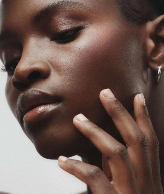 Close-up studio shot of Black woman touching her face. Beauty portrait features skin detail, minimal makeup, silver hoop earrings.