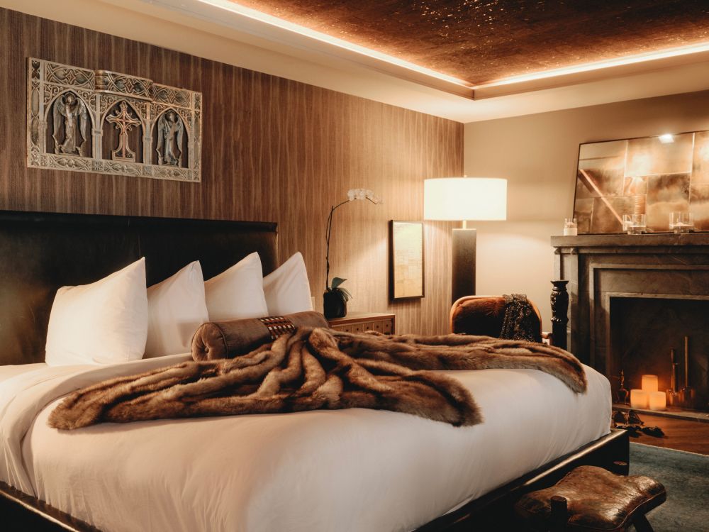 Warm hotel suite featuring a king bed with a fur throw, ornate fireplace, and modern lighting for a cozy and luxurious stay.
