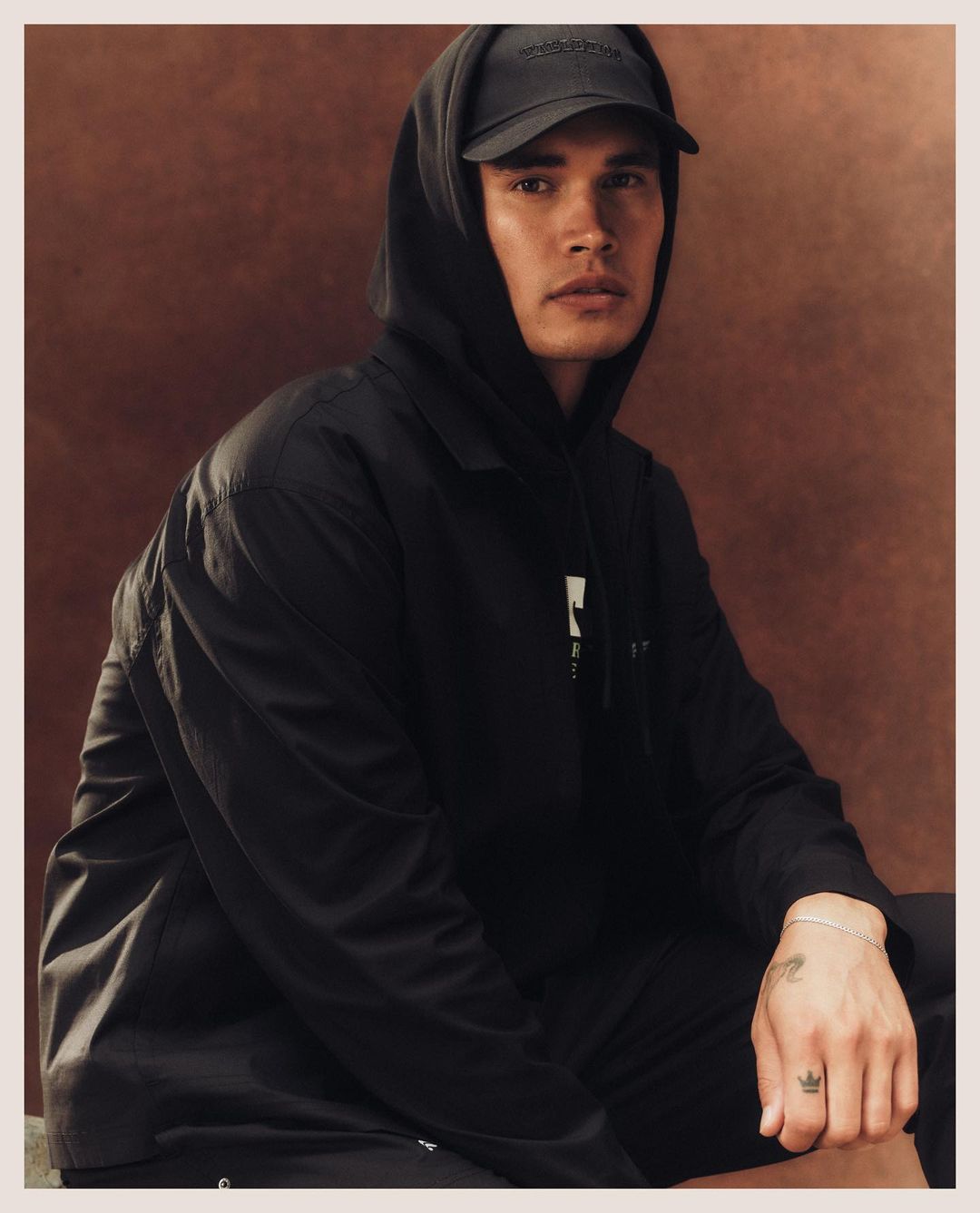 Portrait of an adult male in a black hooded jacket and baseball cap, sitting against a textured brown backdrop, indoor setting.