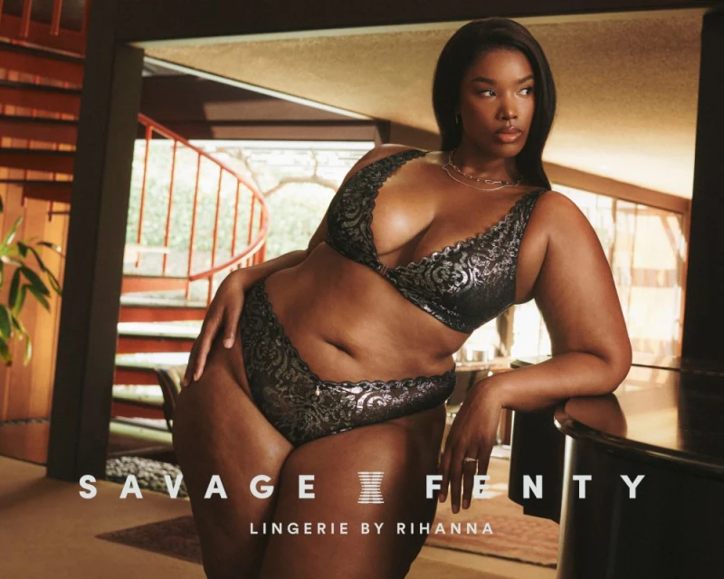 Brunette woman poses in black Savage X Fenty lingerie indoors, showcasing a bra and panty set near a piano and spiral staircase.