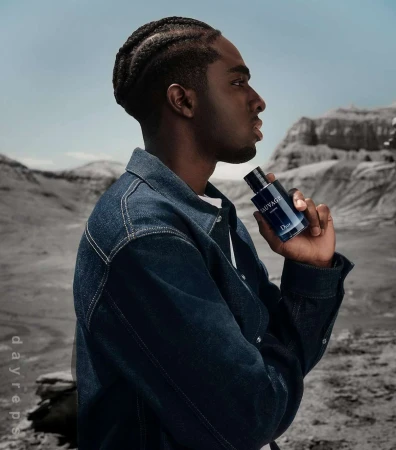 Profile portrait of a dark-skinned adult male wearing a denim jacket and holding a Sauvage Dior cologne bottle in an arid outdoor setting.