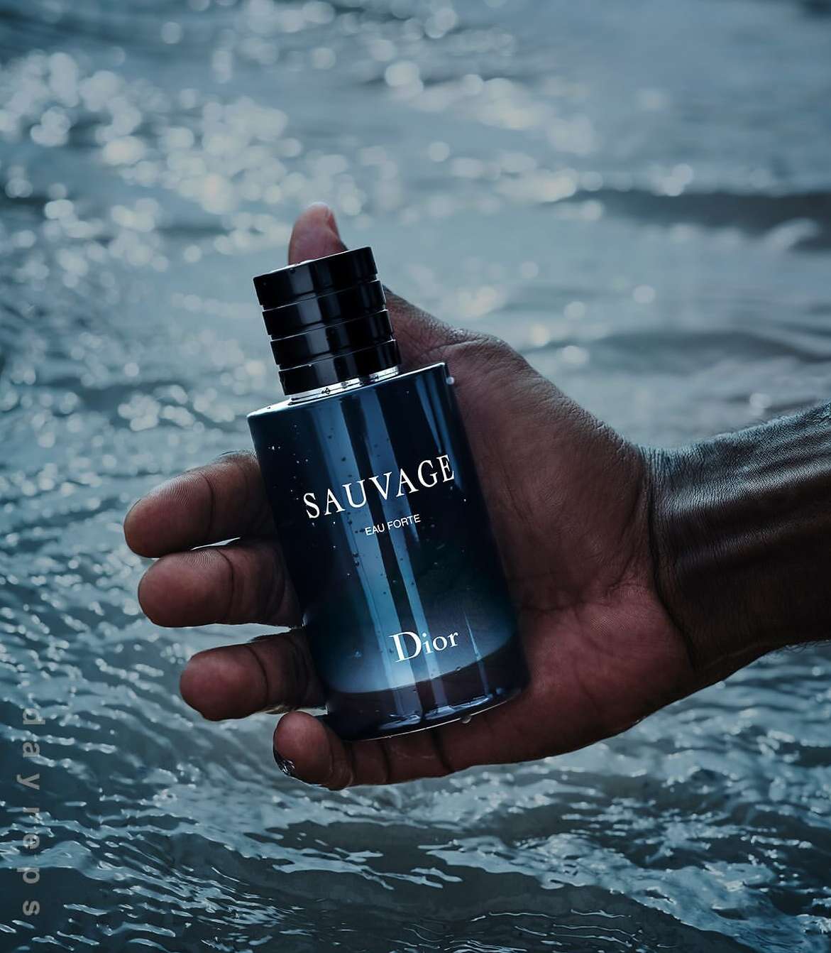 Person holding a bottle of Sauvage Eau Forte cologne by Dior over water. The dark blue bottle is held in a dark-skinned hand.