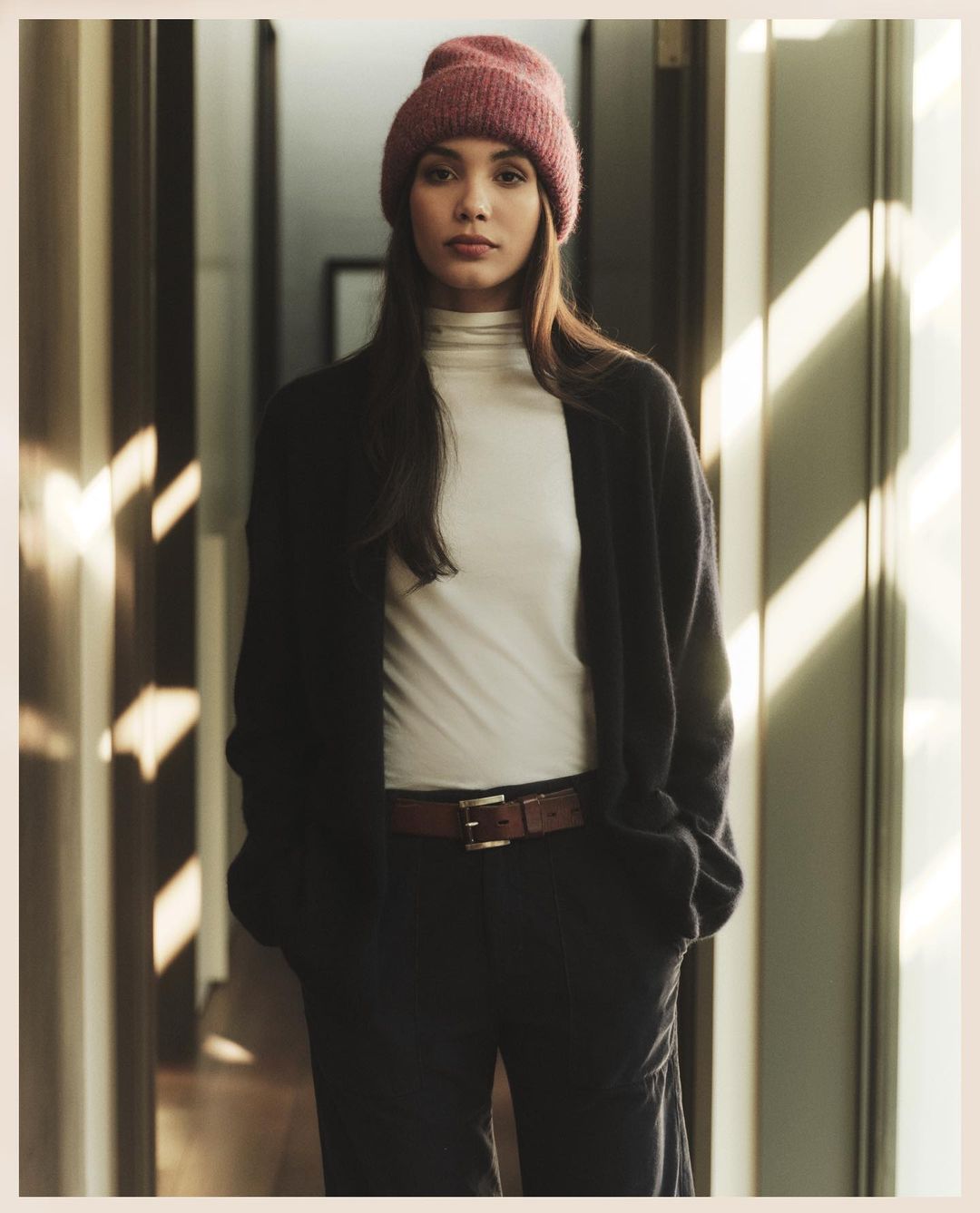 A woman in a pink beanie, white turtleneck, black cardigan, and dark pants stands indoors in natural light by wooden doorways and walls.