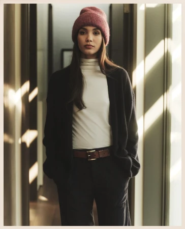 A woman in a pink beanie, white turtleneck, black cardigan, and dark pants stands indoors in natural light by wooden doorways and walls.