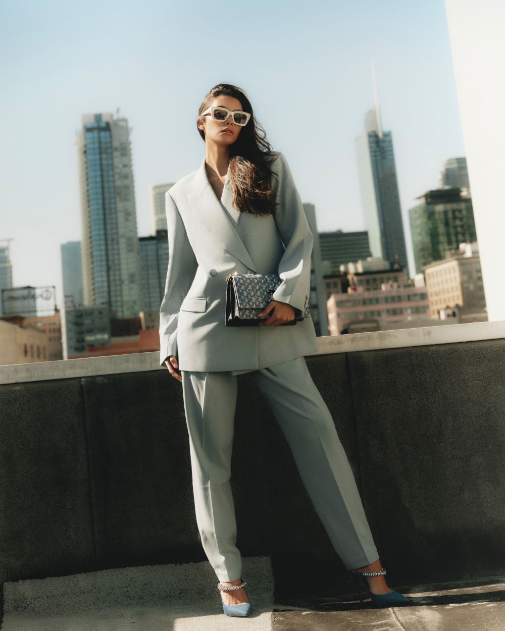 A woman in a light blue pantsuit and sunglasses poses on a rooftop with a handbag, set against a backdrop of city skyscrapers.