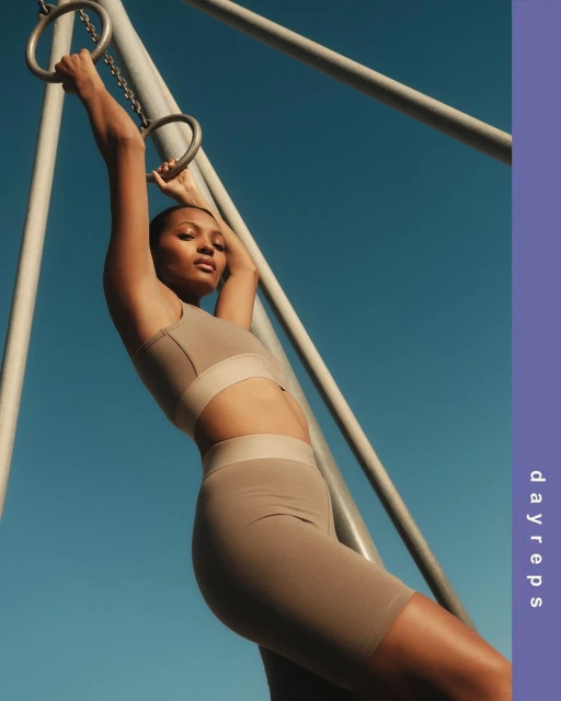 Fit woman in workout gear hangs from metal rings against a blue sky. Athletic female exercising outdoors. Dayreps logo on the right side.