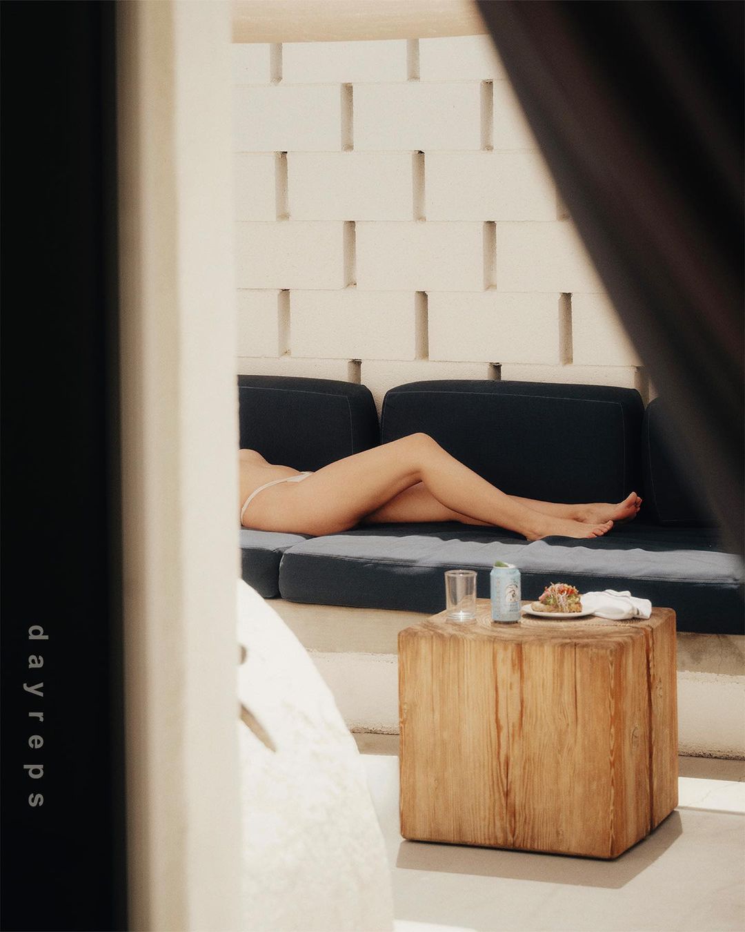 Woman lounges on a navy cushion near a wooden table with a drink and food, framed by geometric white brick and dark curtains.