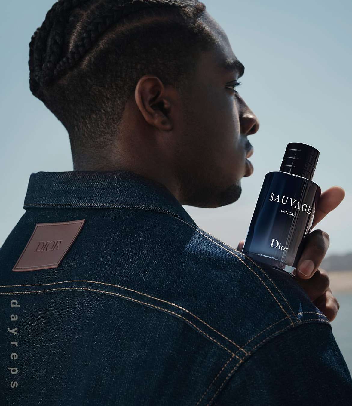 A dark-skinned man in a denim jacket holds a Sauvage Dior cologne bottle against a sky background, showcasing a fragrance ad.
