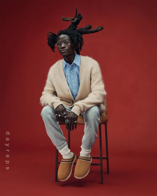 A person with dreadlocks and glasses sits on a stool in a cream cardigan, light blue shirt, and grey pants against a red backdrop.