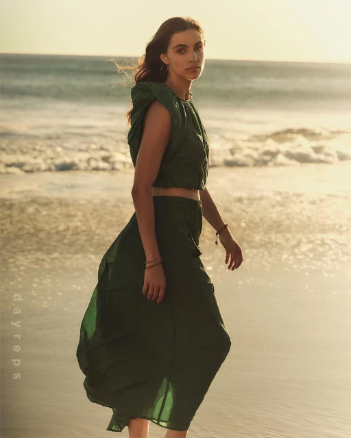 A woman in a green two-piece outfit walks on a sandy beach at sunset, with ocean waves and golden light creating a serene, fashionable scene.
