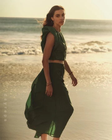 A woman in a green two-piece outfit walks on a sandy beach at sunset, with ocean waves and golden light creating a serene, fashionable scene.