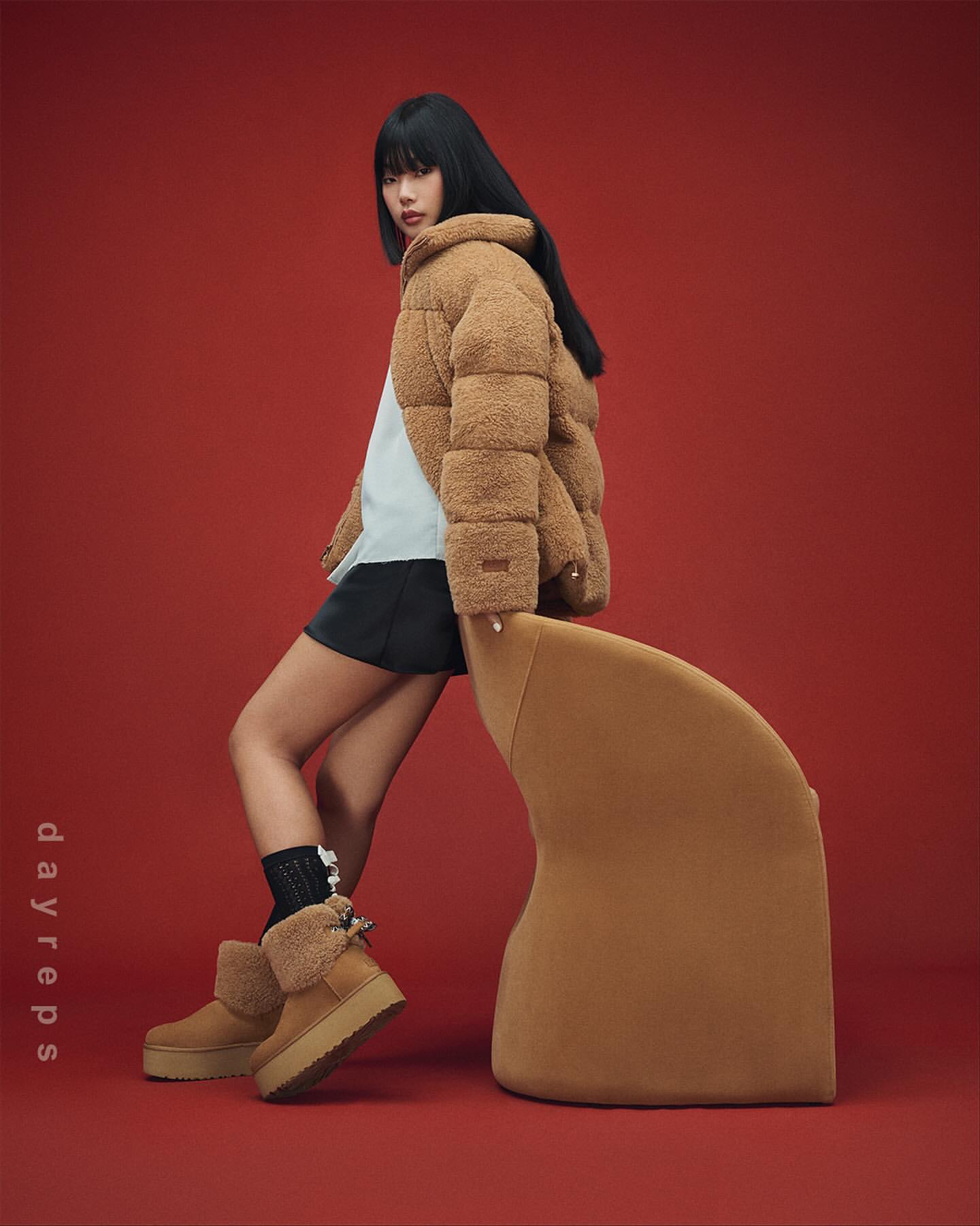 Model in fluffy tan puffer jacket leaning against a matching curved chair on a red studio backdrop