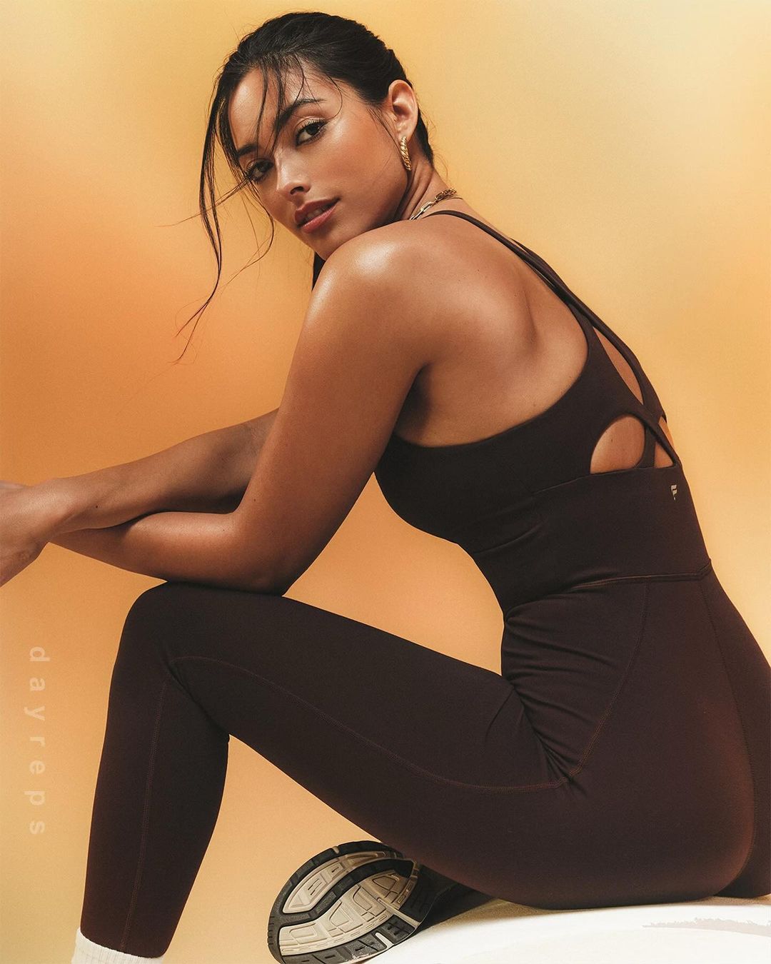 Brunette woman in dark workout clothes poses in a photo studio with soft orange background lighting, looking confidently into the camera.