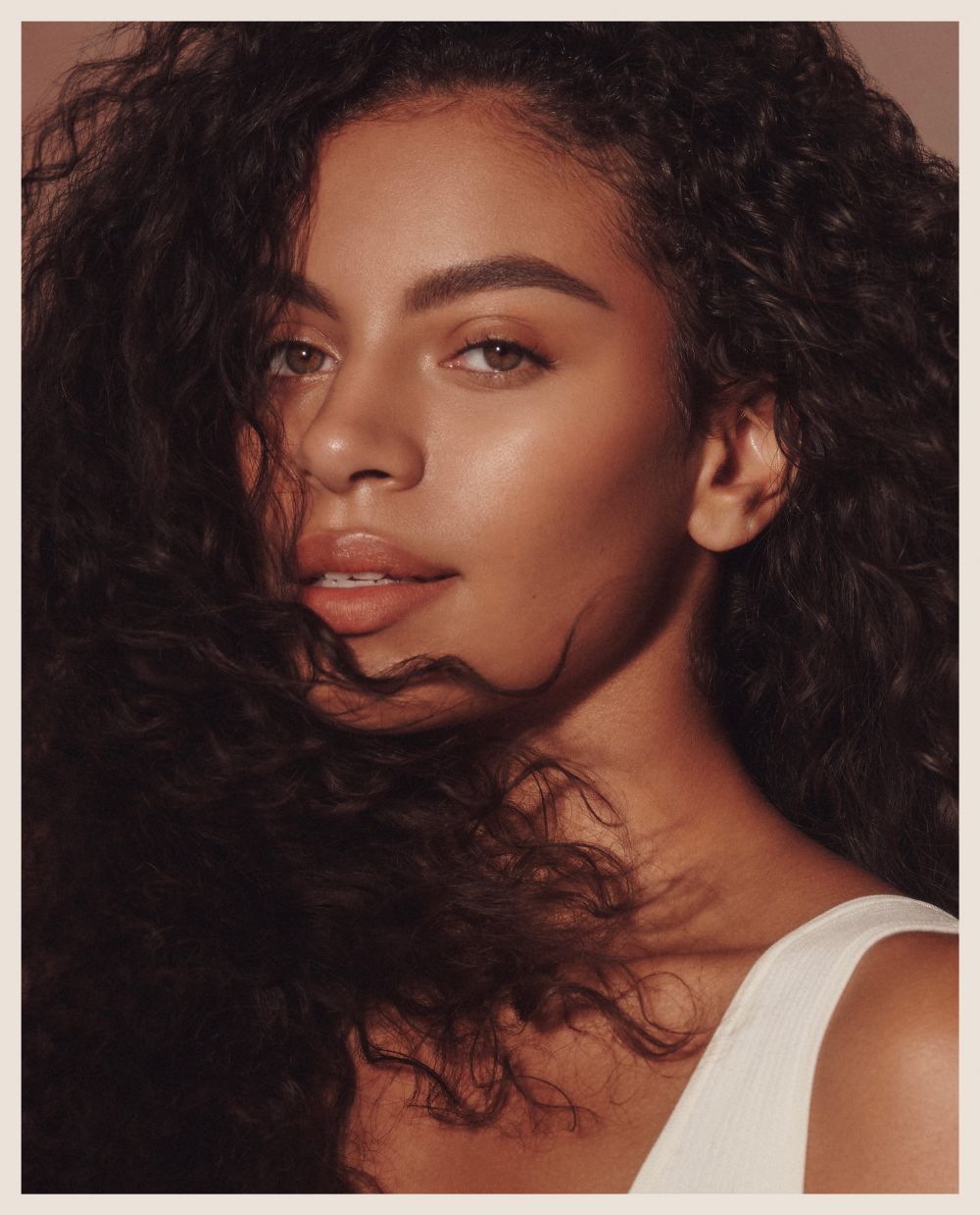 Close-up studio shot of a beautiful woman with curly dark hair, wearing a white top, and softly lit with glowing skin tones.