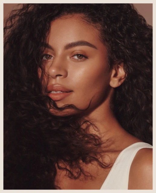 Close-up studio shot of a beautiful woman with curly dark hair, wearing a white top, and softly lit with glowing skin tones.