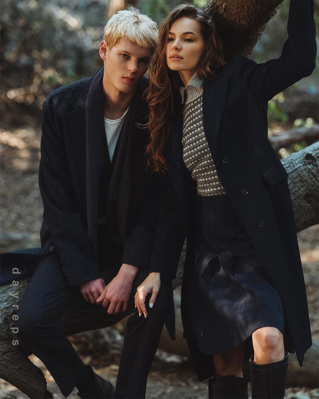 A young blonde man and a brunette woman posing outdoors in a wooded forest setting, wearing dark coats and knitwear.