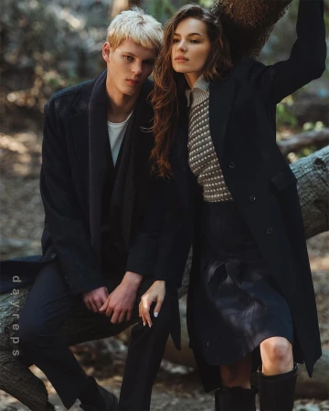 A young blonde man and a brunette woman posing outdoors in a wooded forest setting, wearing dark coats and knitwear.