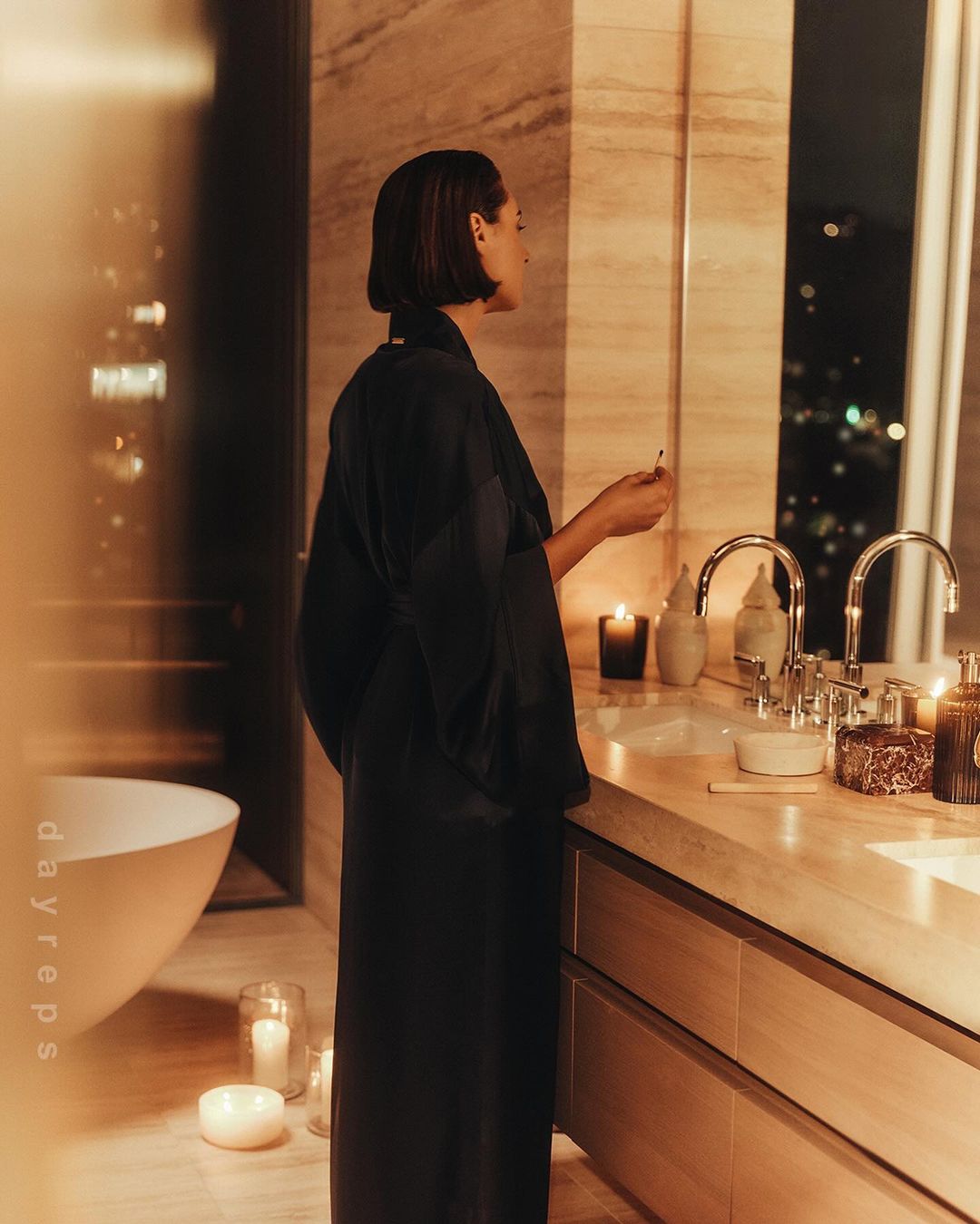 Woman in black robe standing in luxury bathroom with marble vanity, candles, and bathtub; warm lighting and serene atmosphere.