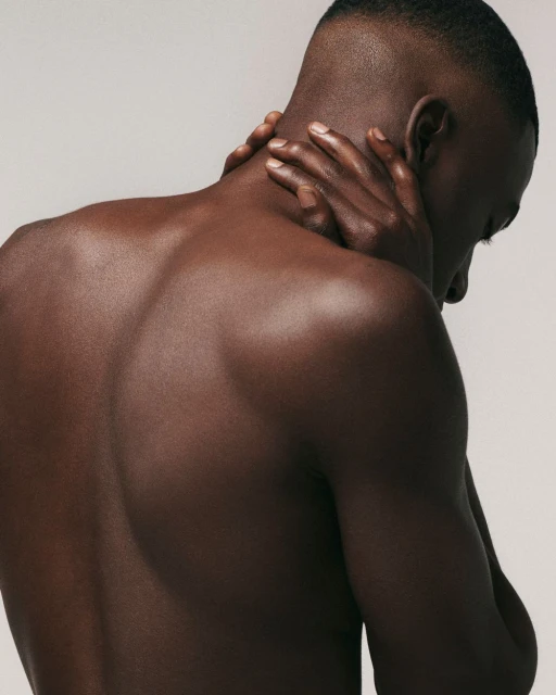 Close-up shot of an adult African American male with short hair, touching his neck with both hands, against a neutral, light-colored background.