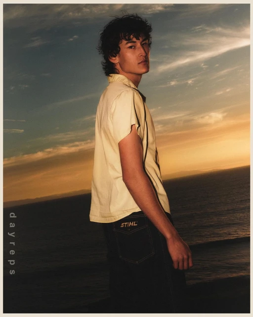 Portrait of a dark-haired man in a light yellow shirt and dark jeans with a logo on the pocket, posing outdoors near the ocean.