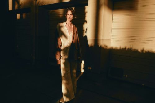 Woman in a light suit stands indoors with patterned shadows, posing against roll-up doors in warm, diffused light. Fashion editorial.