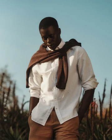 Black man wearing a white button-down shirt and brown trousers with a brown sweater draped over his shoulders outdoors