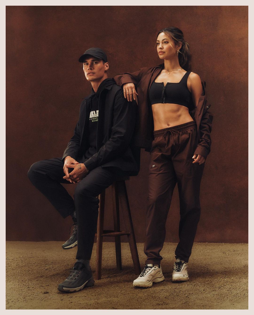 Man seated on stool next to standing woman wearing brown jacket and black crop top against textured warm backdrop