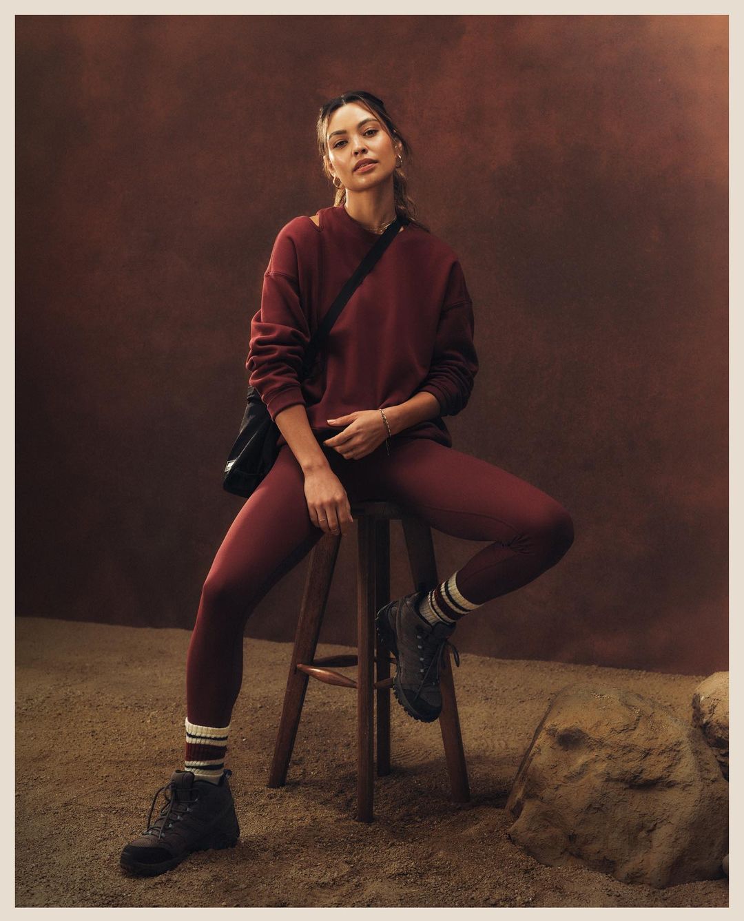 Woman in a maroon sweatshirt and leggings sitting on a wooden stool in a sandy environment, wearing black boots and carrying a black bag.