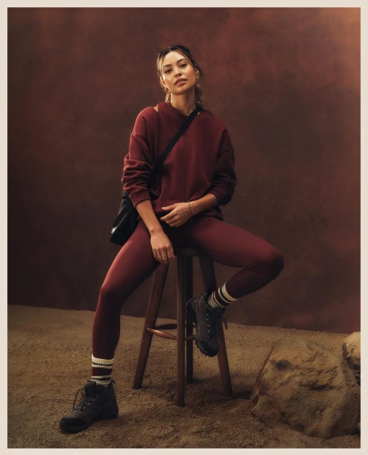 Woman in a maroon sweatshirt and leggings sitting on a wooden stool in a sandy environment, wearing black boots and carrying a black bag.