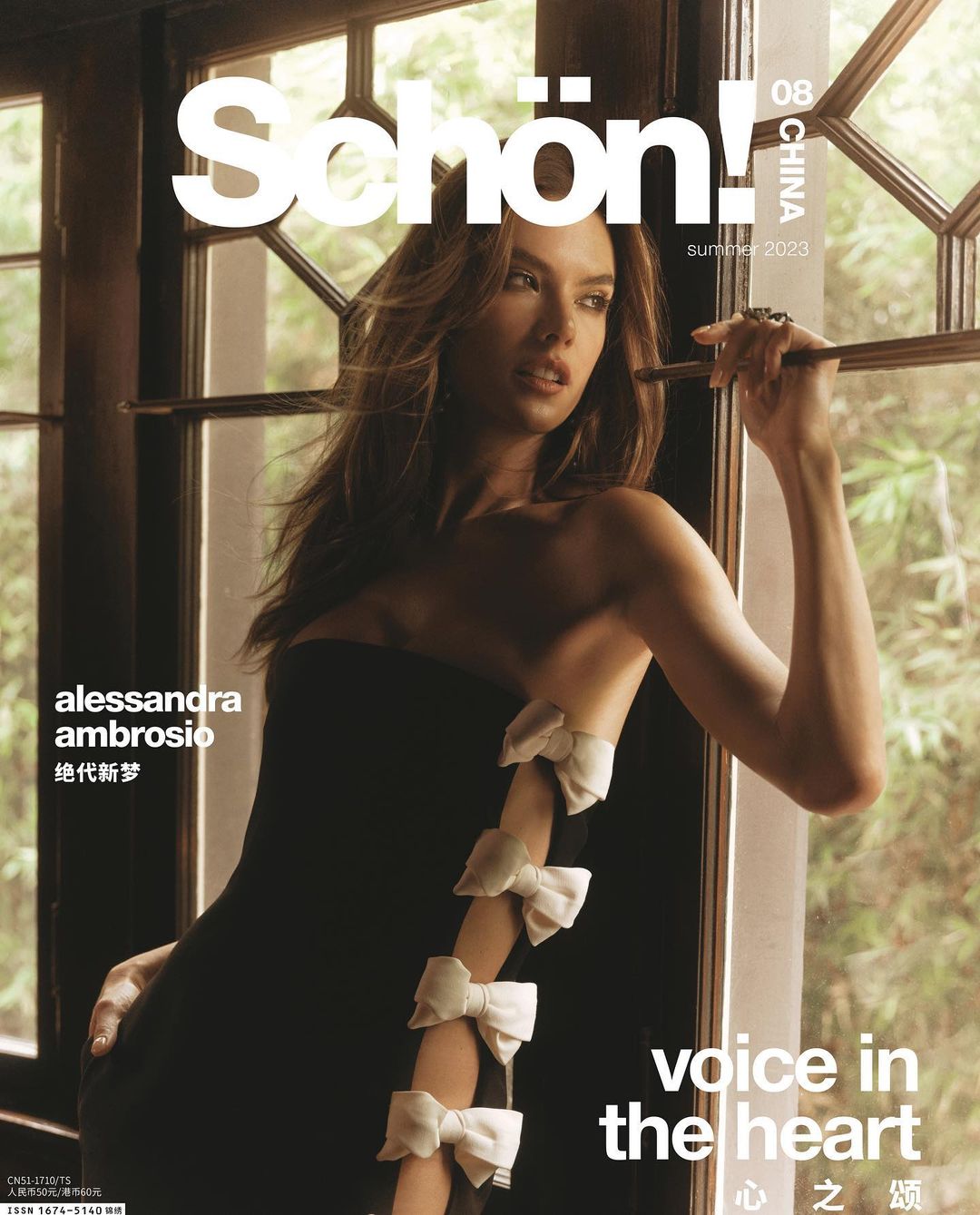 Alessandra Ambrosio on the cover of Schön! China Summer 2023 magazine, wearing a black strapless dress adorned with white bows by a window.