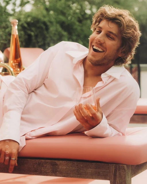Blonde man lounges on an outdoor bed, smiling and holding a drink. The background features green plants, adding a fresh touch.