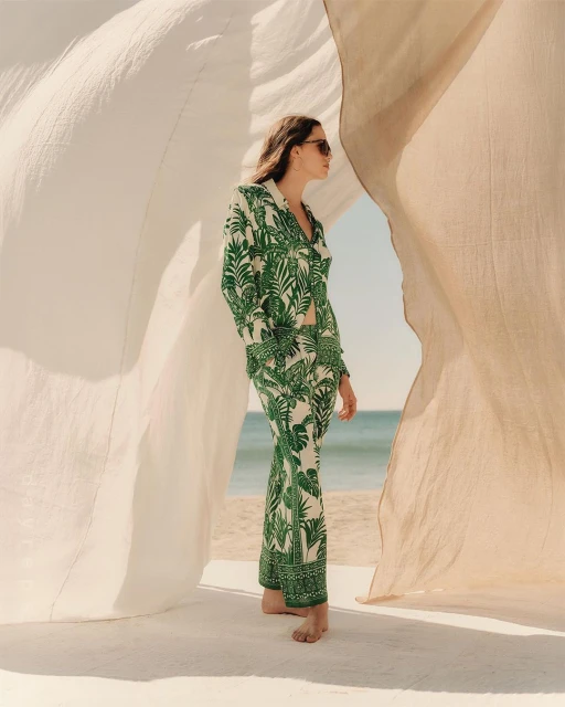 Woman in a green and white tropical print suit standing on a beach with white draped fabric and blue water in the background.