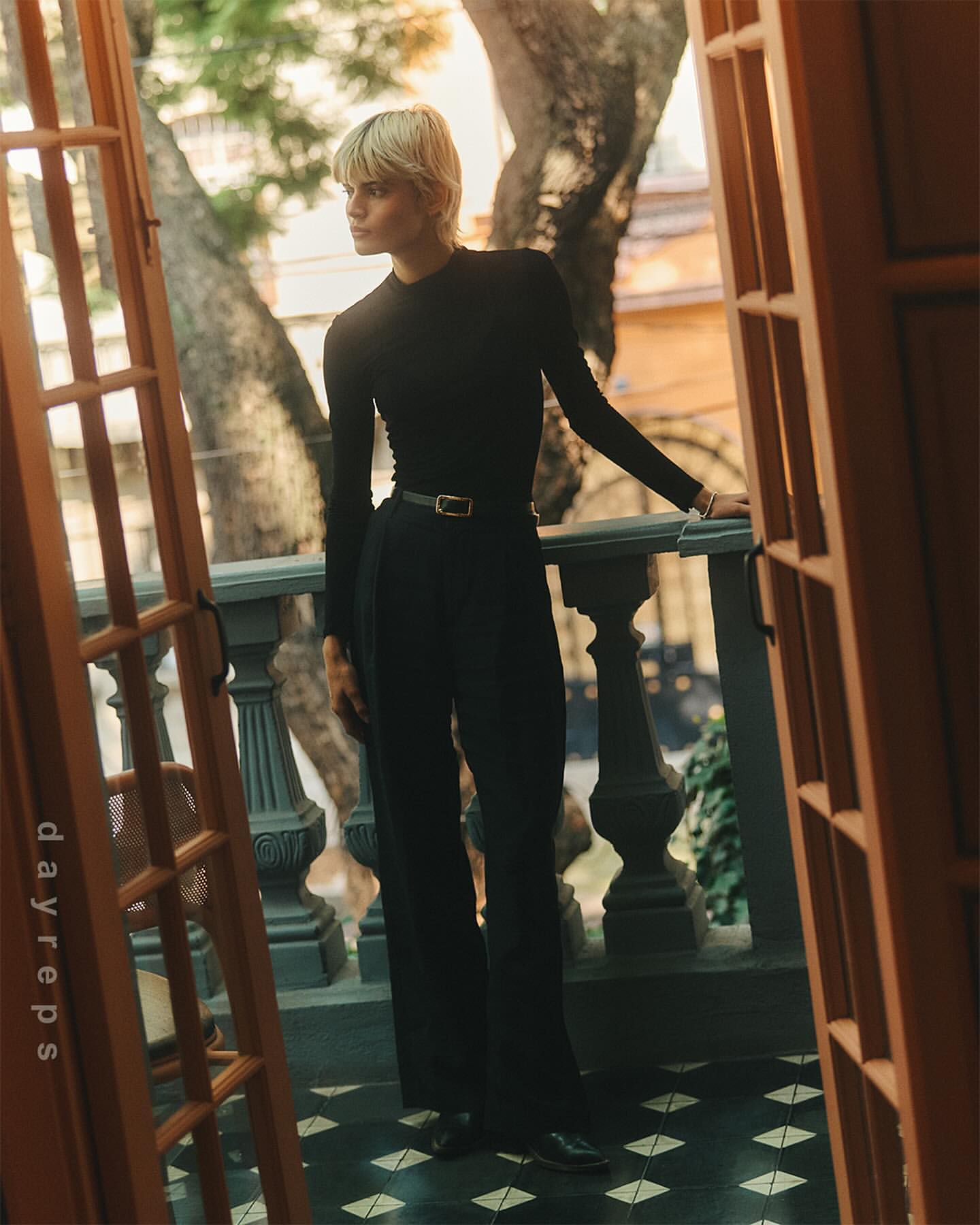 Blonde woman in black long sleeve and pants standing on a balcony with geometric floor tiles, framed by a partially open wooden door.