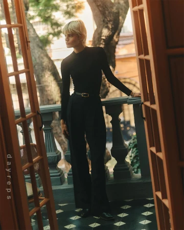 Blonde woman in black long sleeve and pants standing on a balcony with geometric floor tiles, framed by a partially open wooden door.