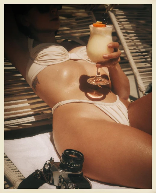 Woman in bikini lying on hammock holding cocktail with camera on towel, suggestive of vacation, relaxation and tropical travel photography.