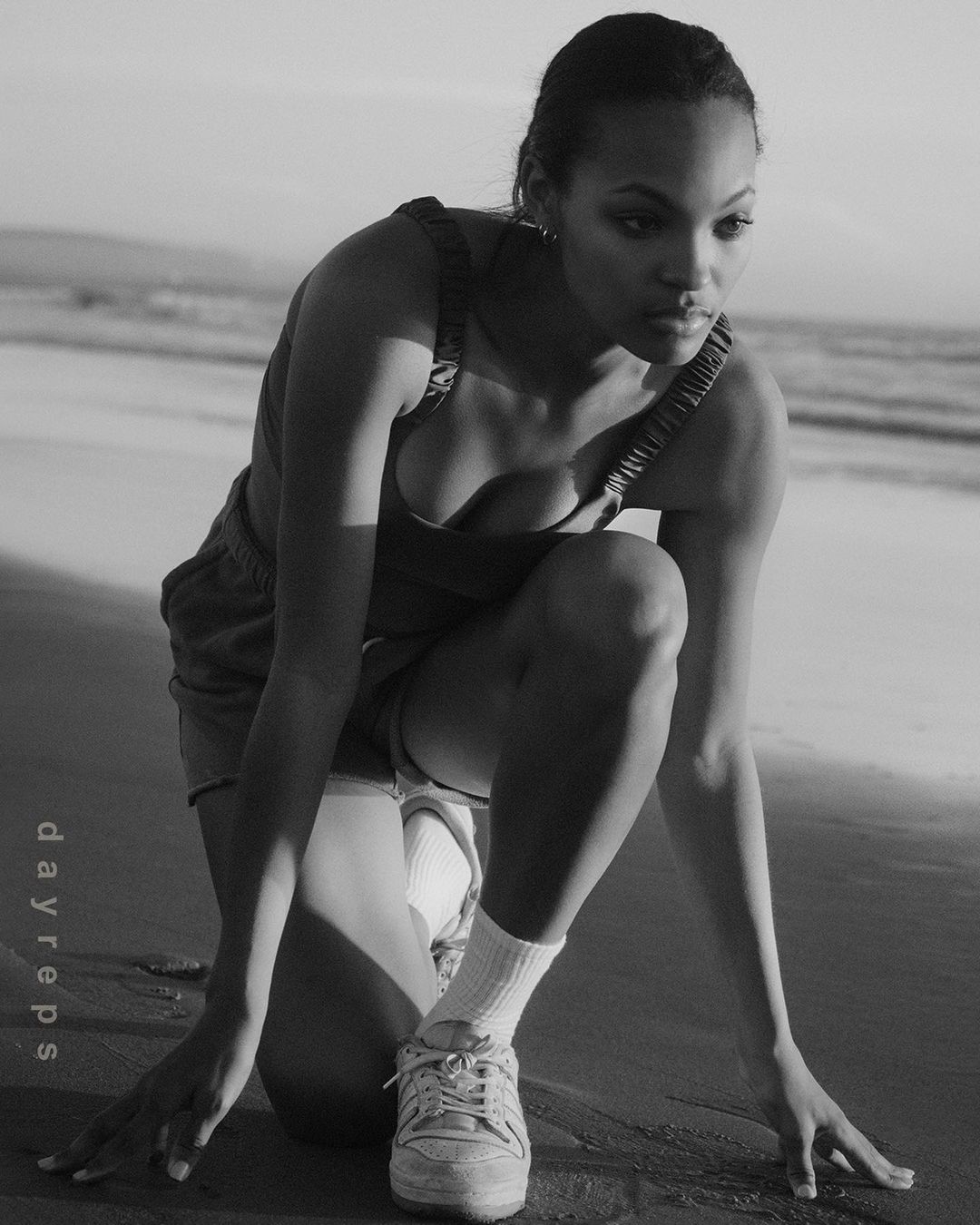 Black and white portrait of a woman in workout clothes kneeling on a sandy beach, gazing intently at something out of frame.