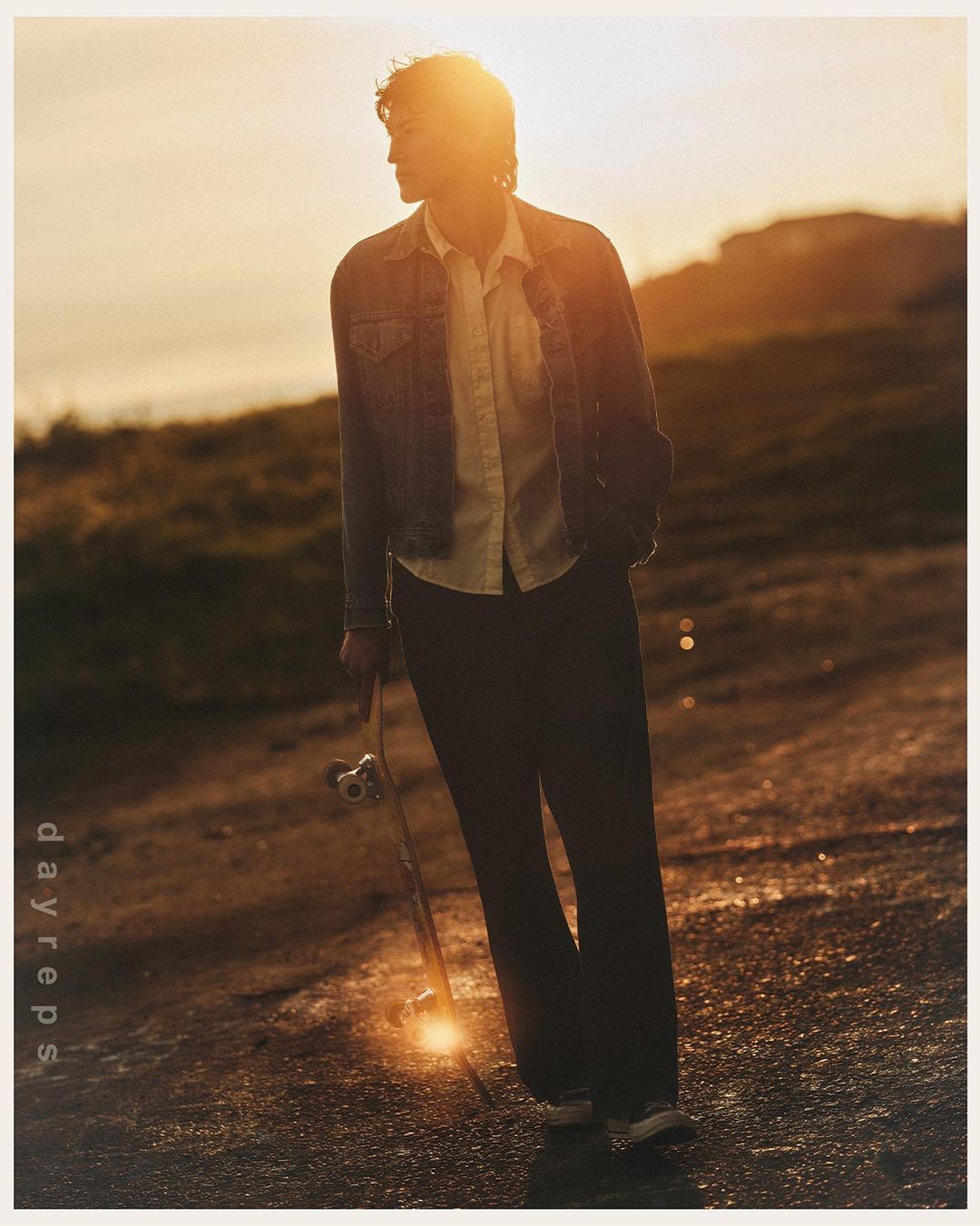 A man wearing a denim jacket and holding a skateboard standing outdoors during sunset.