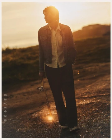 A man wearing a denim jacket and holding a skateboard standing outdoors during sunset.
