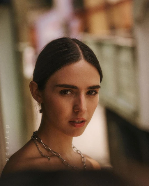 Close-up portrait of a woman with dark hair and light skin, wearing silver chain jewelry, soft natural lighting in an indoor setting.
