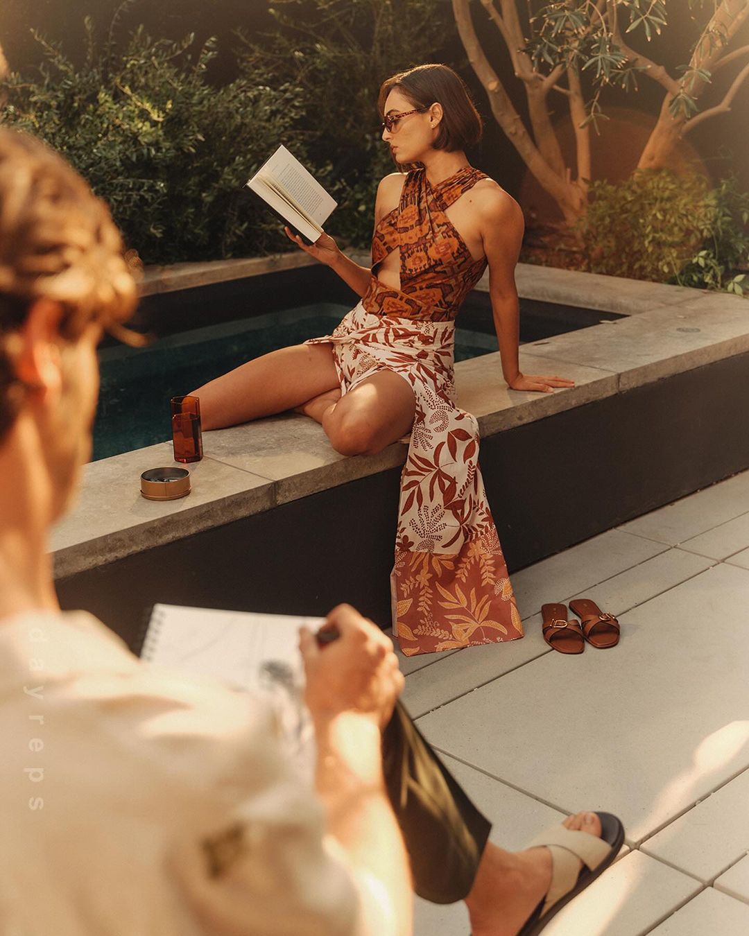 Woman in floral-print swimsuit and wrap sits poolside reading a book while a man sketches her portrait outdoors in natural light.