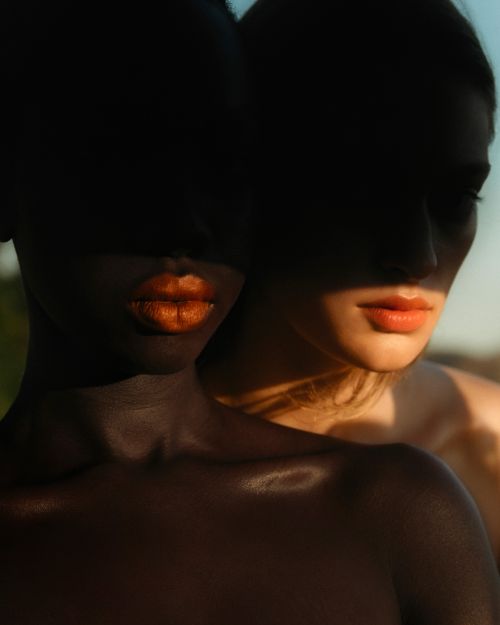 Two women with dramatic lighting, one with dark skin and orange lipstick, the other with fair skin and matching lipstick outdoors.