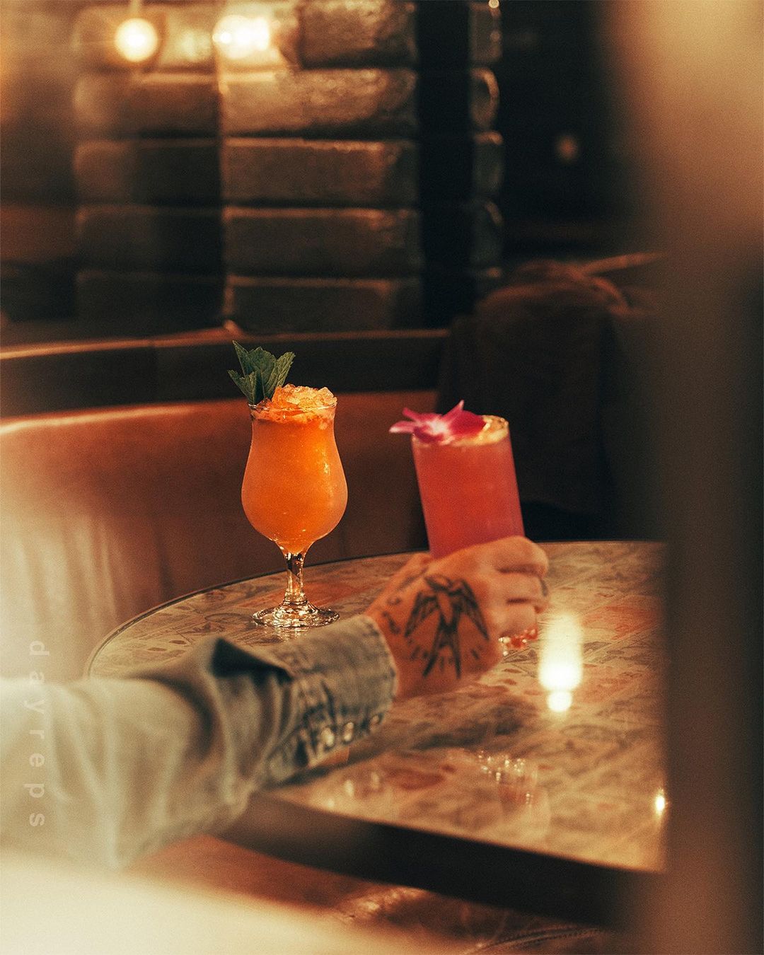 Two cocktails on a marble table, one orange and one pink, with a hand holding the pink drink in a dimly lit bar or restaurant.