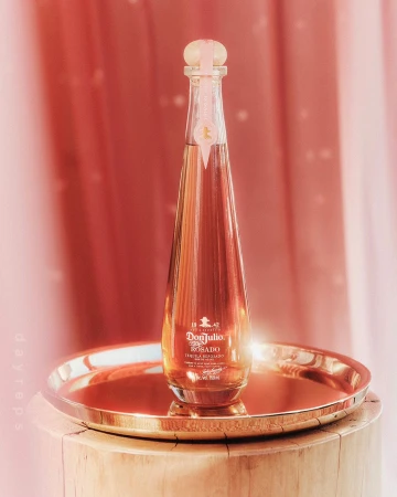 Elegant bottle of Don Julio Rosado tequila on a gold tray atop a rustic wooden stool, set against a soft pink backdrop. Studio shot.