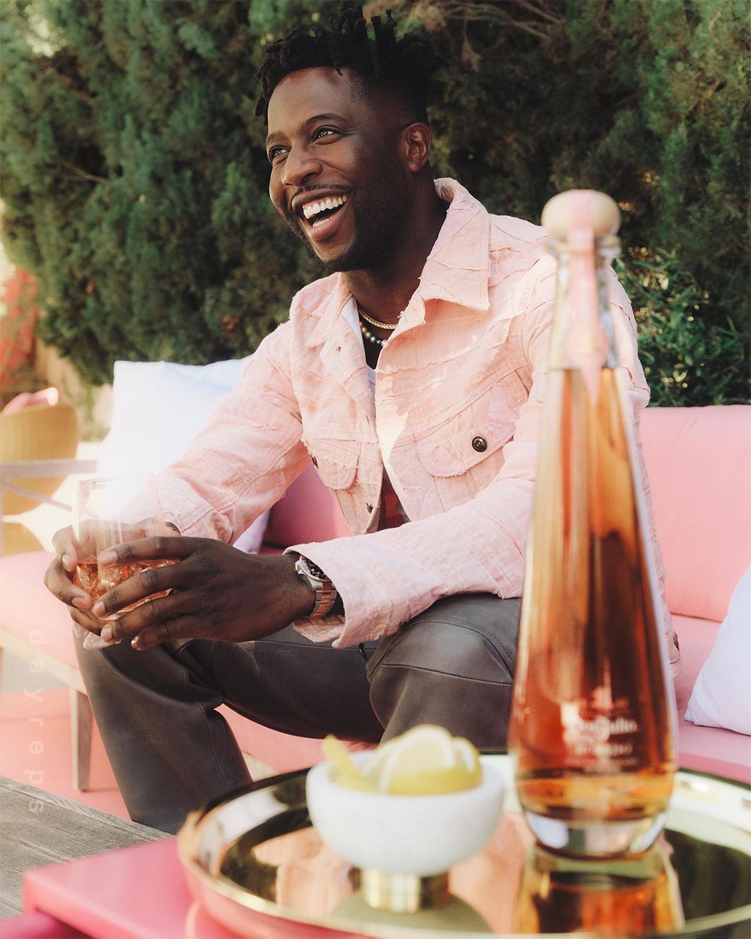 Smiling African-American man in pink jacket seated outdoors with drink; bottle, lemon slices, green foliage background. Sunny, relaxed setting.