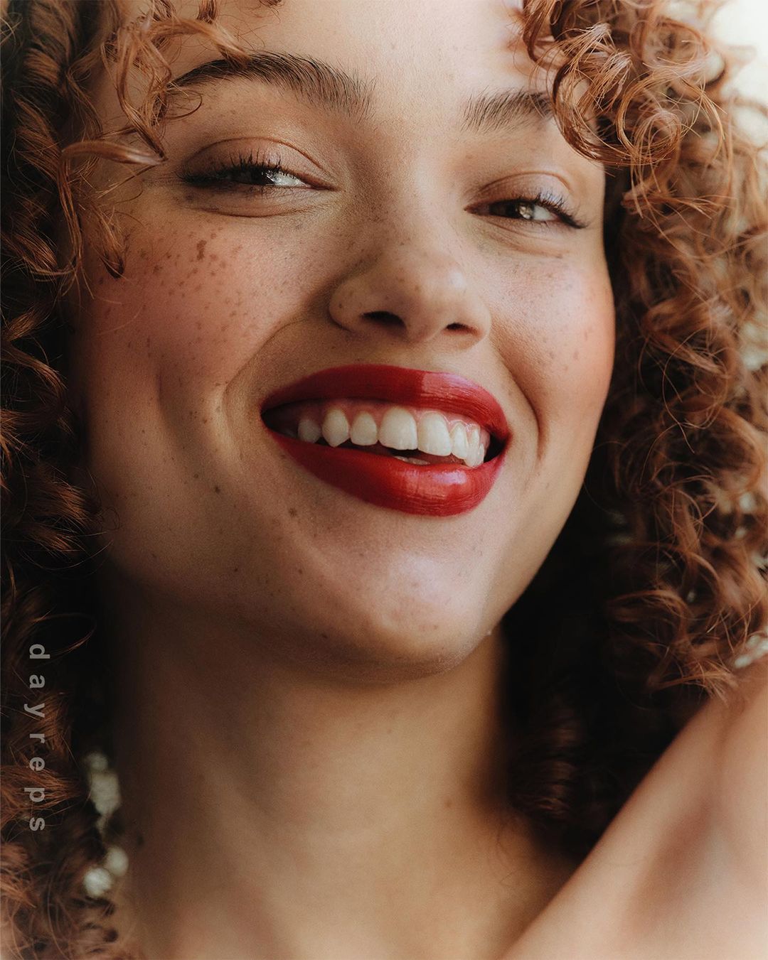 Close-up portrait of a smiling young woman with curly red hair, freckles, and glossy red lipstick photographed by Wolfe + Von.
