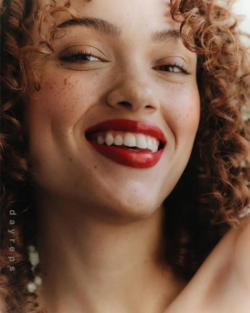 Close-up portrait of a smiling young woman with curly red hair, freckles, and glossy red lipstick photographed by Wolfe + Von.